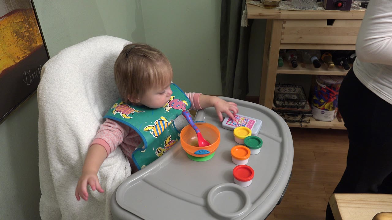 Baby in a high chair reaching toward a toy phone and colorful cups.