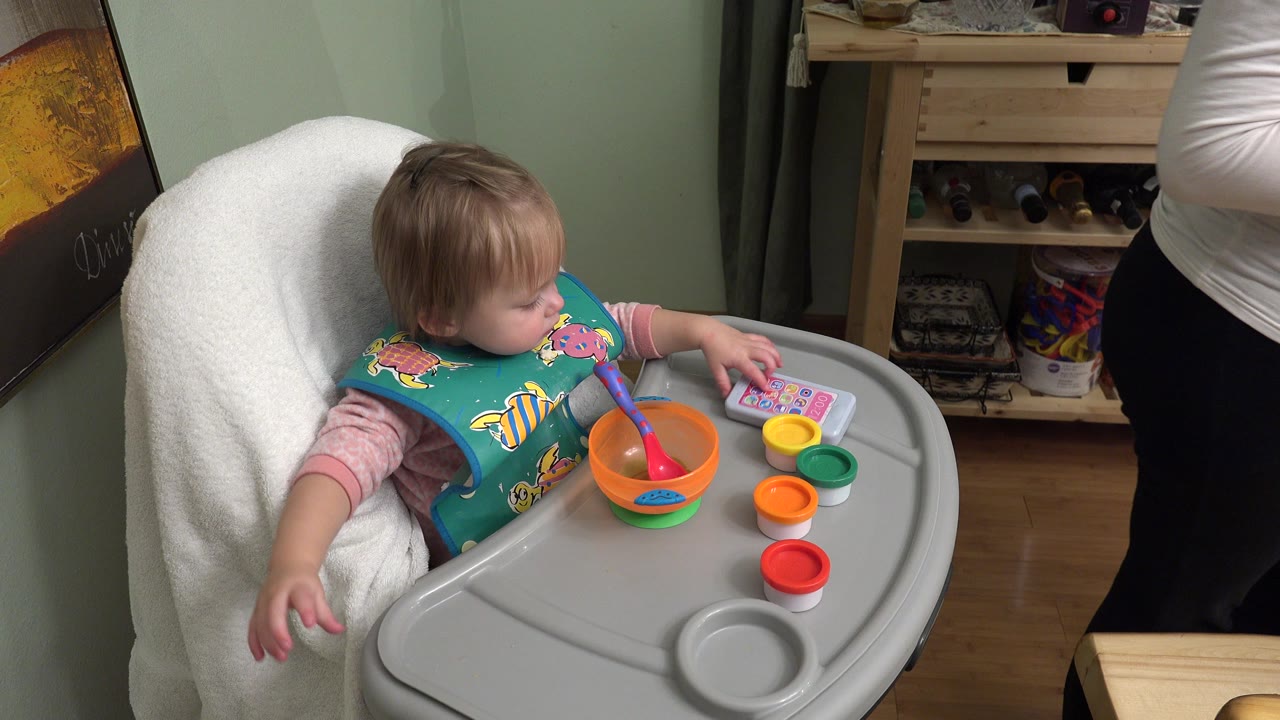 The Westminster, Colorado home movie frame shows the baby looking down at the tray, with an orange bowl, spoon, colored cups, and toy phone arranged in front.