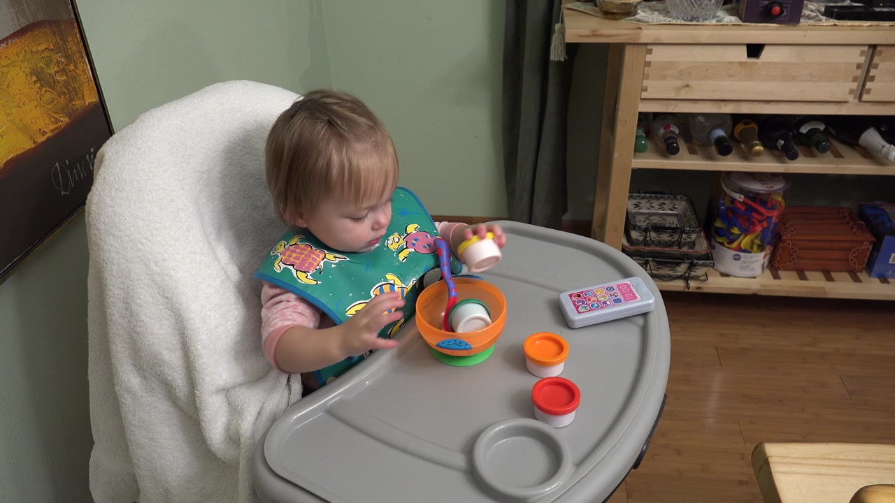 Blonde toddler in a gray high chair reaching toward an orange bowl.