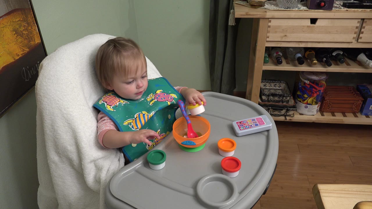 Baby in a high chair reaching toward colorful toys on the tray.