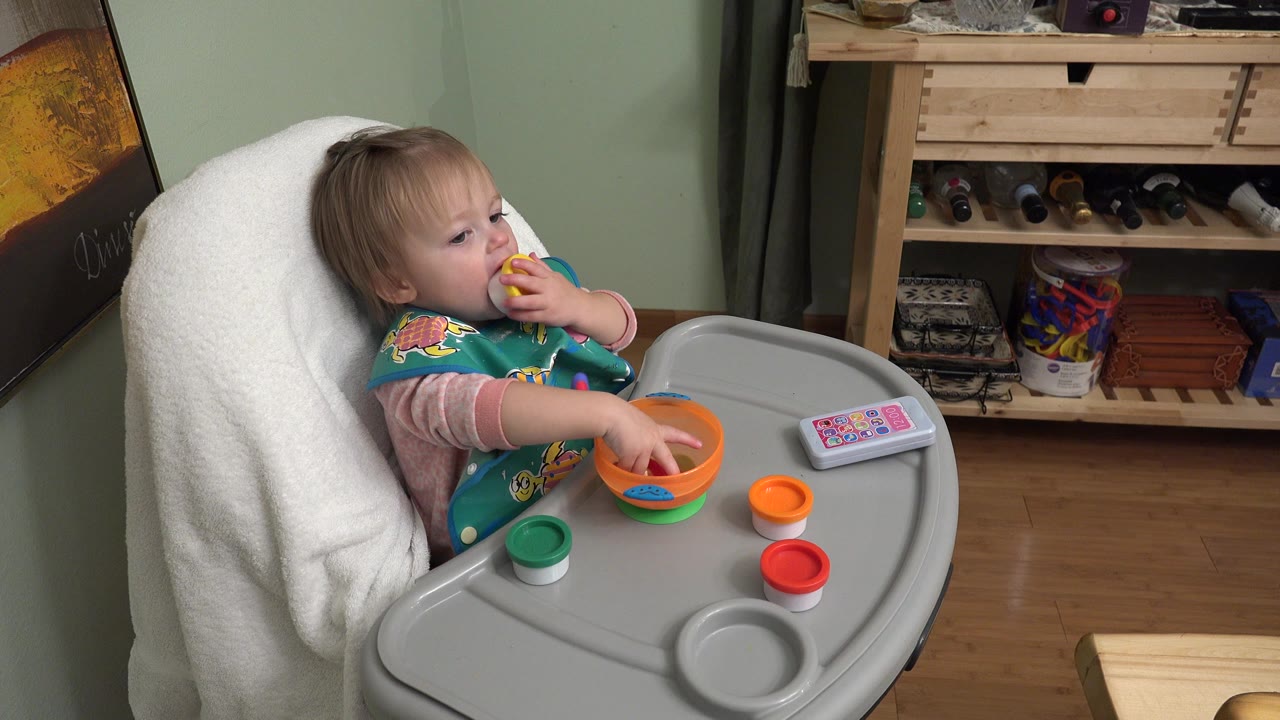 Baby in a meal chair holding food near her mouth beside an orange bowl.