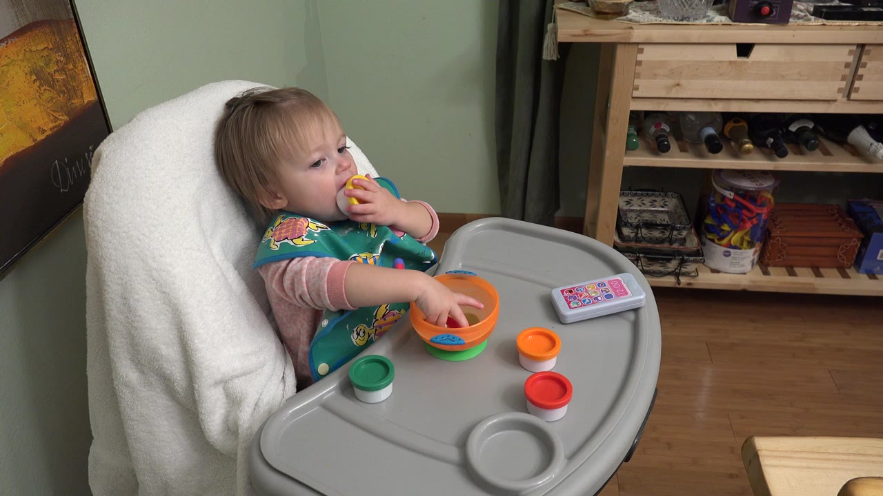 At 0:01 in Westminster, Colorado, the baby remains seated in the high chair, looking toward the tray while holding the yellow food and keeping one hand near the orange bowl.