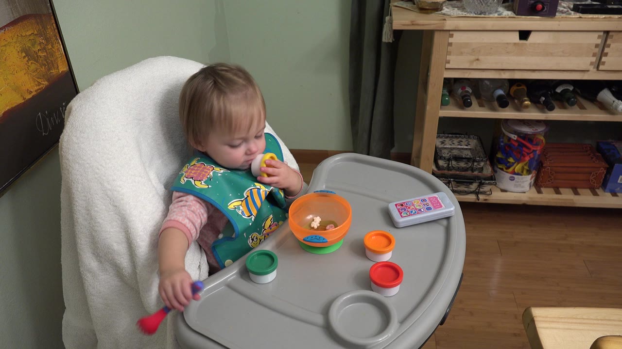 Baby in a highchair holding toys beside an orange bowl.
