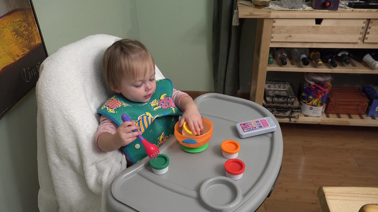 At 0:01, the toddler looks down toward the tray in the Westminster home interior, with the red utensil near a green cup and an orange bowl close to the right hand.