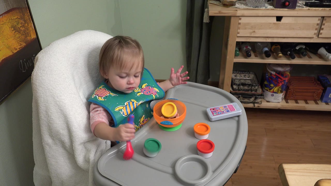 Baby in a high chair holding a red spoon near colorful toy cups.