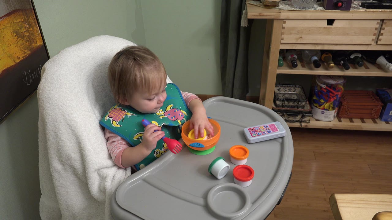 Toddler in a high chair holding toy utensils over a gray tray.