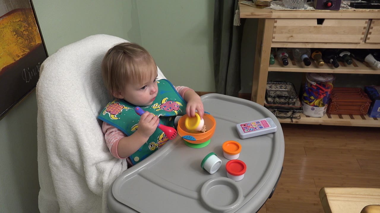 Blond baby in a high chair holding toy utensils beside an orange bowl.