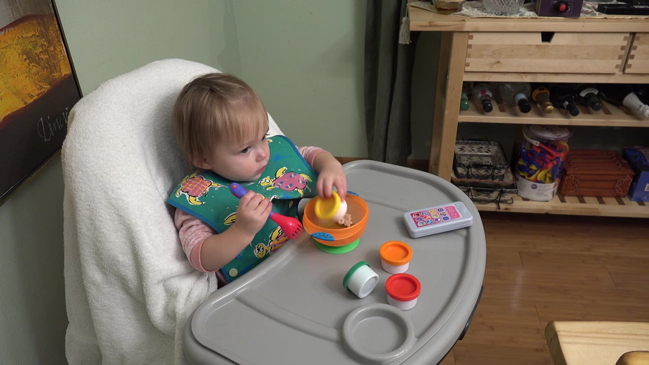 In Westminster, Colorado, the baby looks to the side while holding a yellow-and-white toy pot above the orange bowl on the high chair tray.