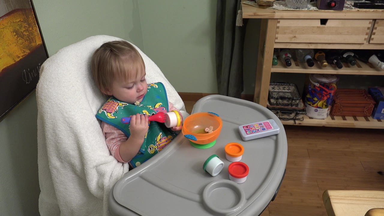 Toddler in a highchair holding a red spoon beside an orange bowl.