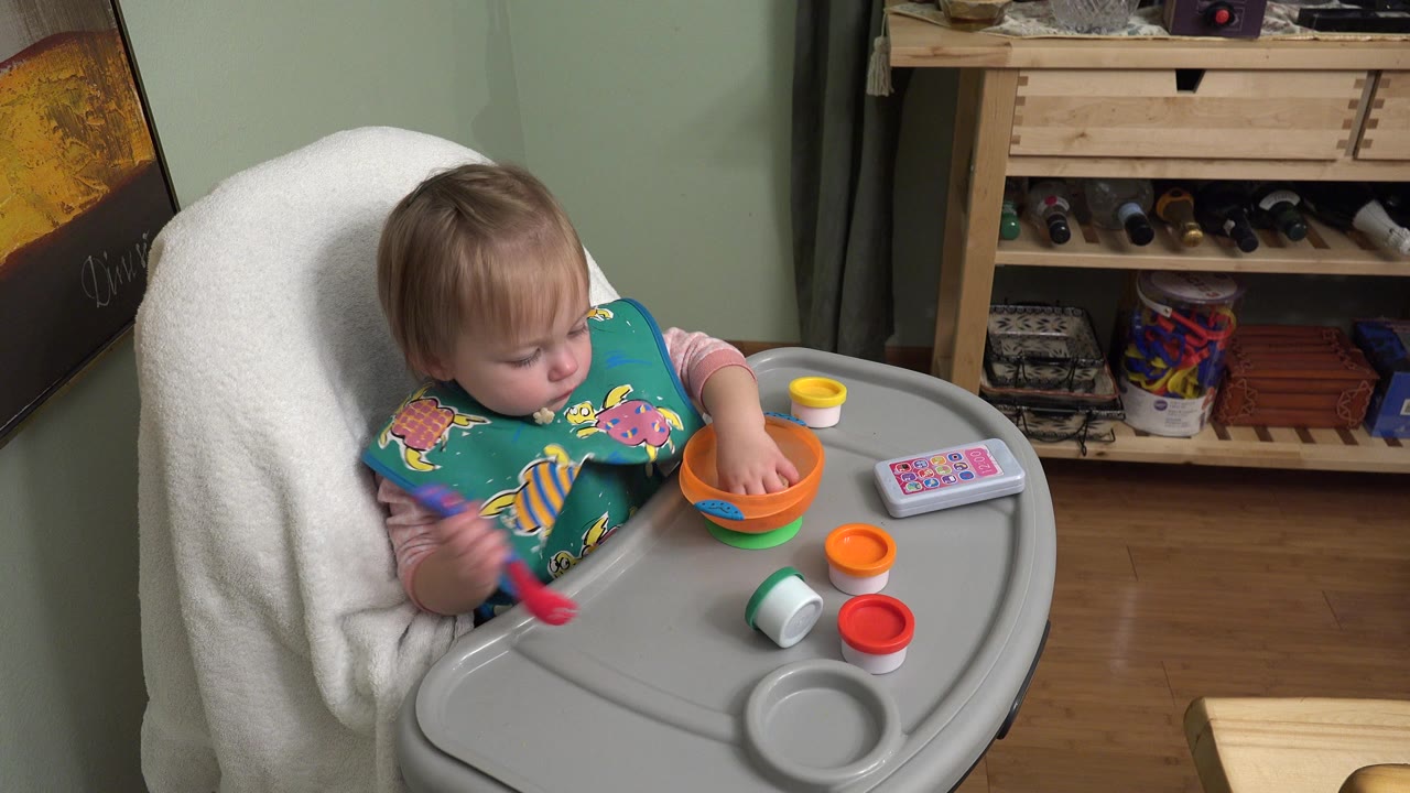 In Westminster, Colorado, a baby in a highchair looks down at an orange bowl while a red spoon, toy lids, and a small toy phone sit on the tray.