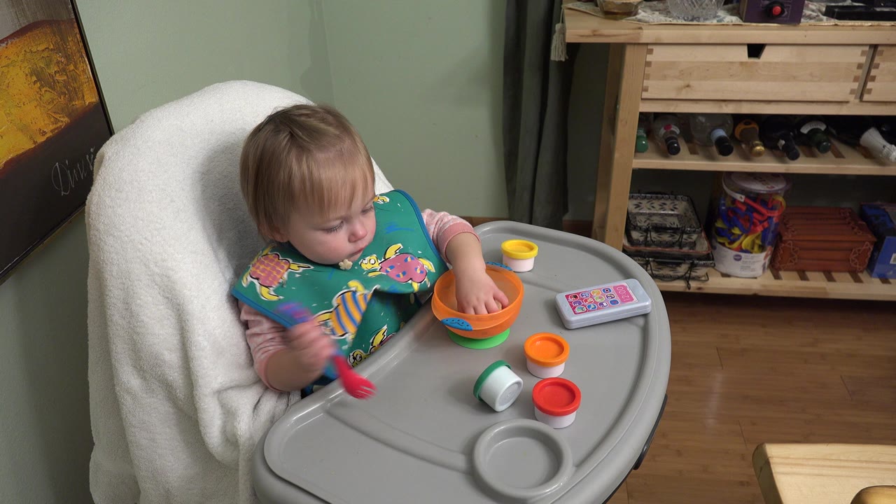 In Westminster, Colorado, the baby holds a red spoon near the tray and studies the orange bowl, with a toy phone and colorful lids arranged to the right.