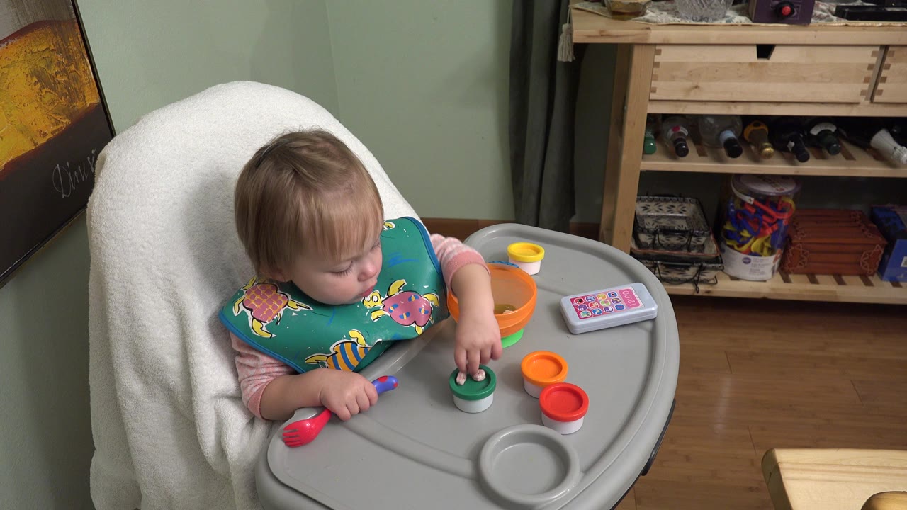 Baby in a high chair reaching toward a green toy cup.