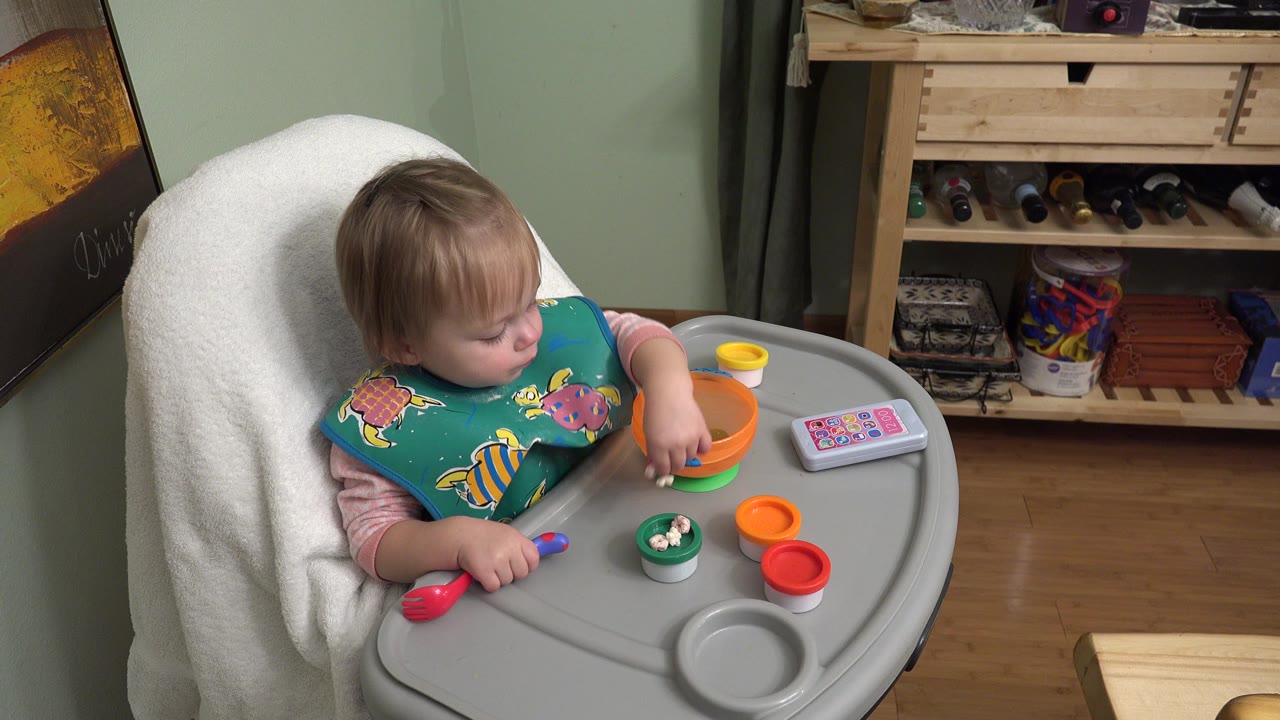 Toddler in a green smock sitting in a grey highchair with small paint containers on the tray.