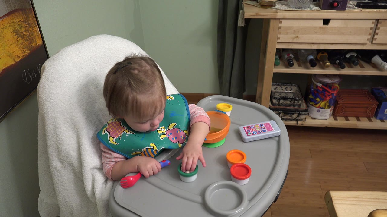 Blond baby in a gray baby chair reaching toward colorful toy pieces.