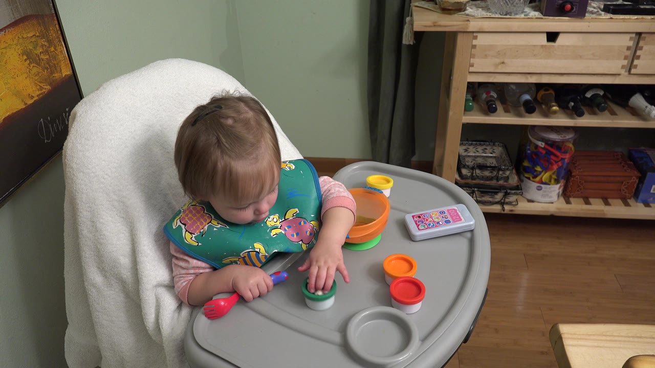 The baby remains seated in the Westminster home interior, looking down while one hand rests near a green cup and other colorful pieces on the gray tray.