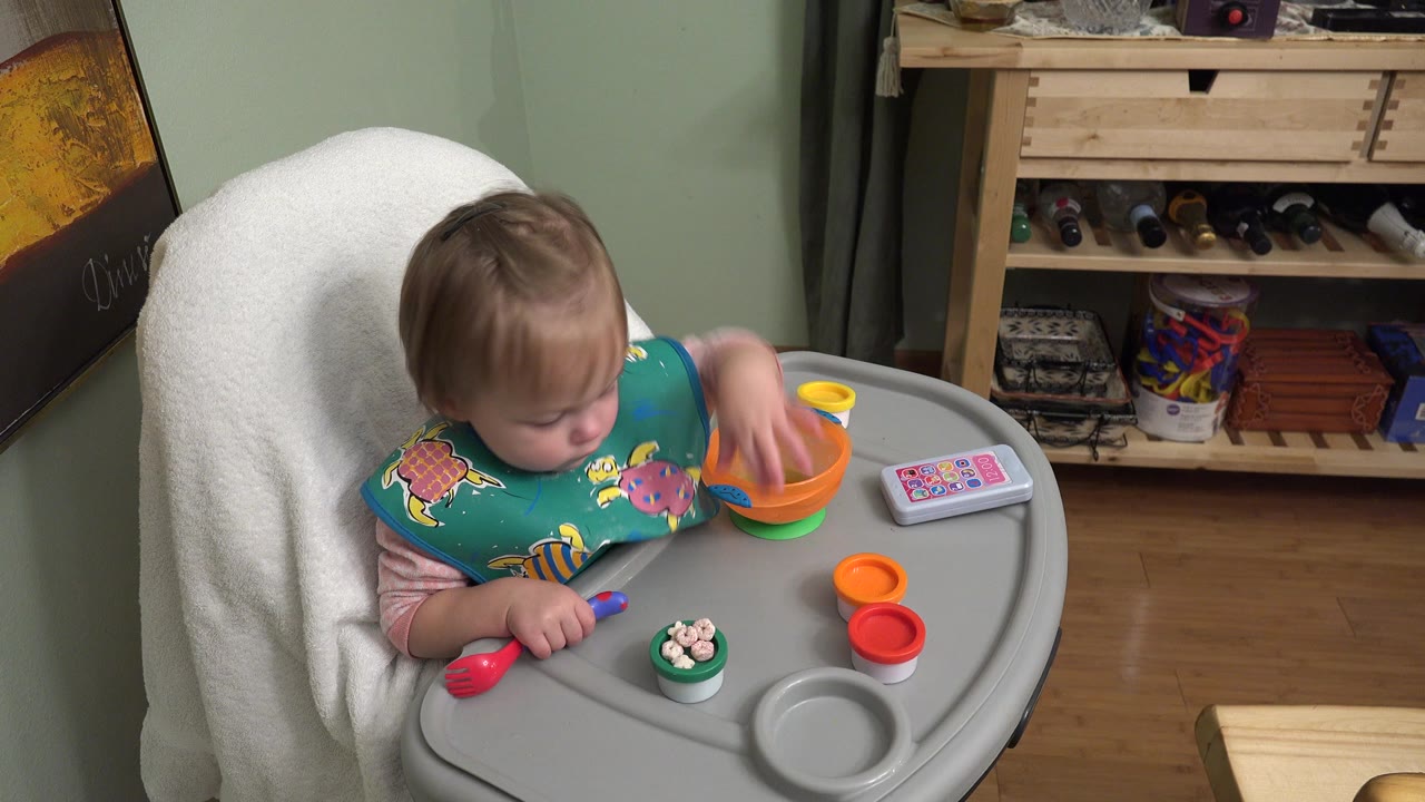 In Westminster, Colorado, the toddler keeps one hand inside an orange bowl while toy spoons, toy food, and a small rectangular toy sit on the high-chair tray.