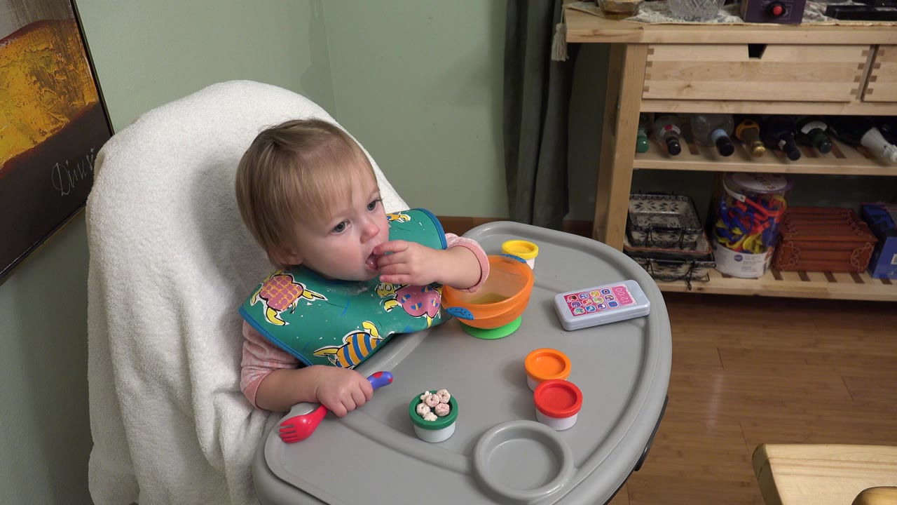 Baby girl in a high chair eating finger food beside colorful toys.