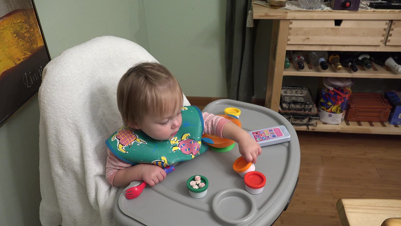 Blond baby in a chair reaching toward orange and red toys on a tray.