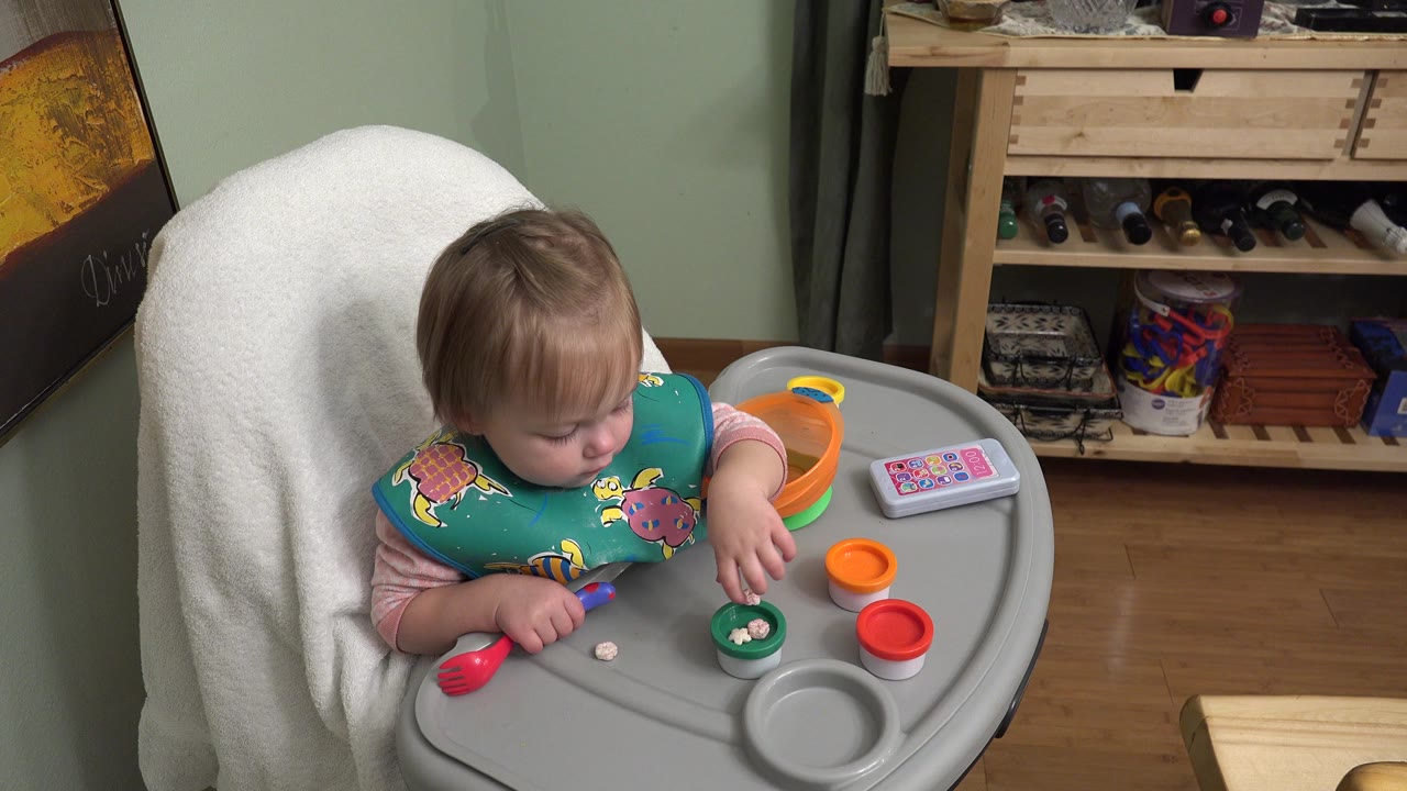 At 0:01 in Westminster, Colorado, the child leans over the high-chair tray with one hand near a green cup containing small food pieces.