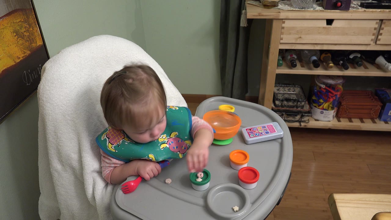 Young child in a high chair reaching toward small toys on a gray tray.