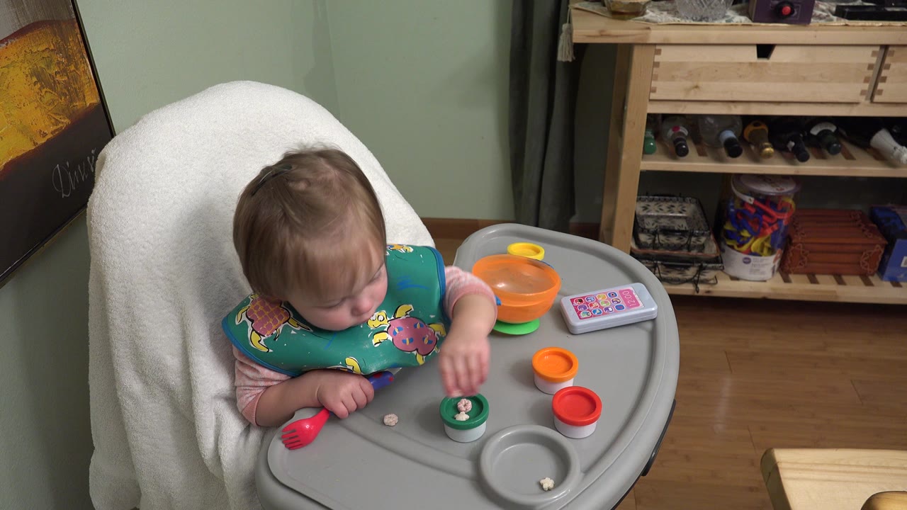 At 0:01 in Westminster, Colorado, the child leans forward over the high-chair tray with one hand near a green toy cup and the other near a red utensil.