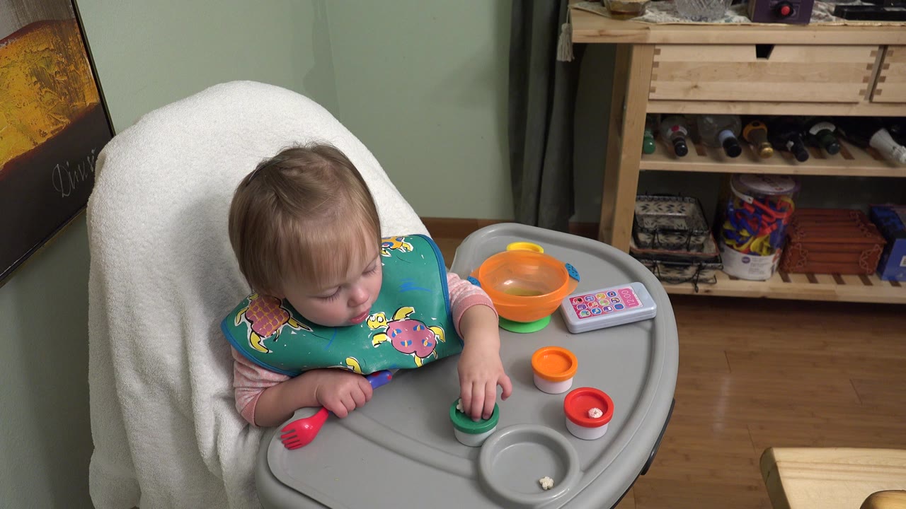 Young girl in a high chair looking down at a tray with bowls and a red utensil.