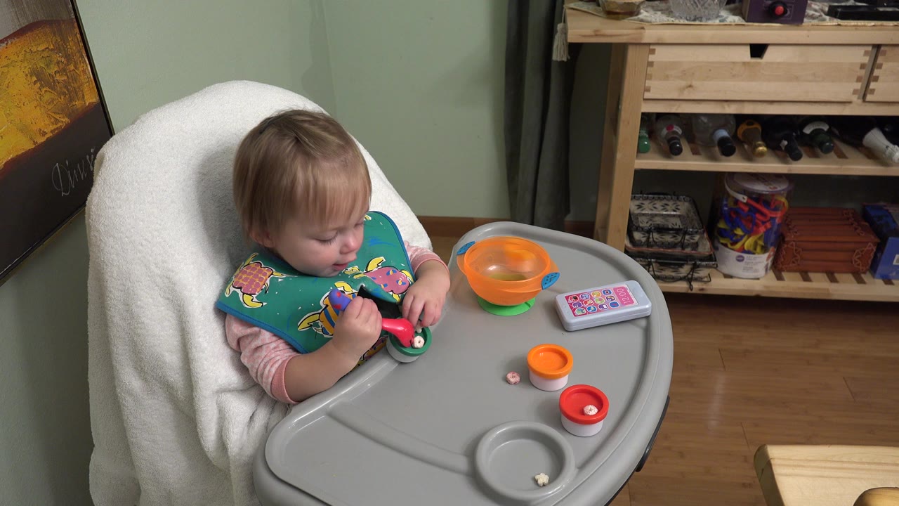 Toddler girl in a high chair holding a toy beside an orange bowl.