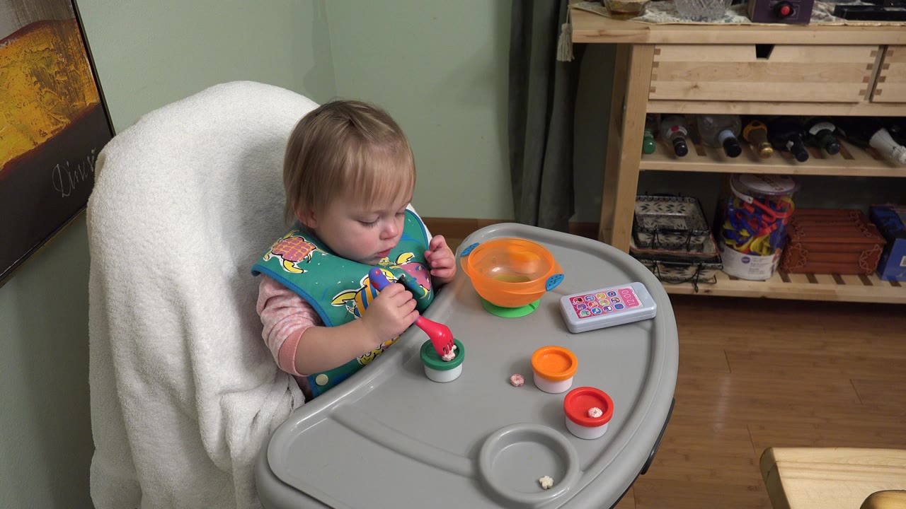 At 0:00 in Westminster, Colorado, a young child sits in a small chair, looking down at a red and green spoon near an orange bowl on a gray tray.