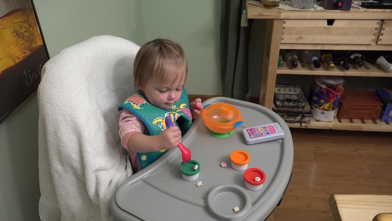 Toddler in a high chair touches a small cup on a tray.
