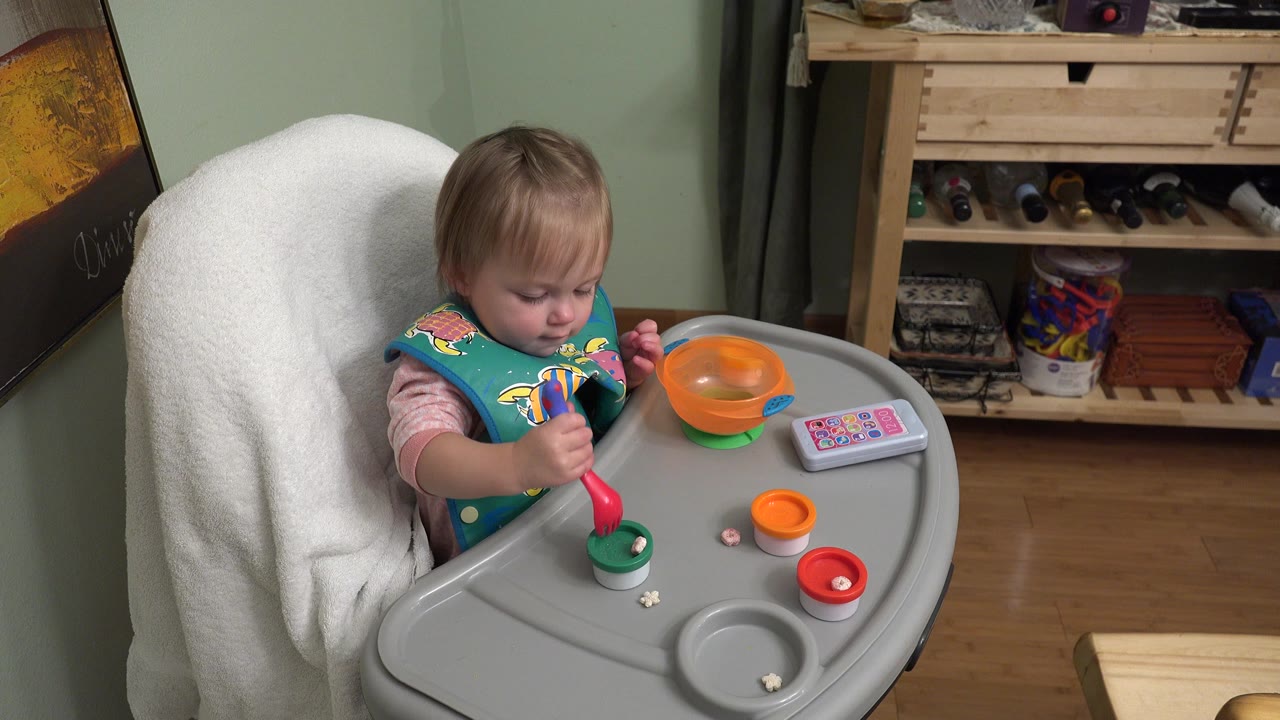 A toddler in Westminster, Colorado looks down at the high-chair tray while holding a red spoon near a small cup, with an orange bowl and colorful small objects spread across the surface.