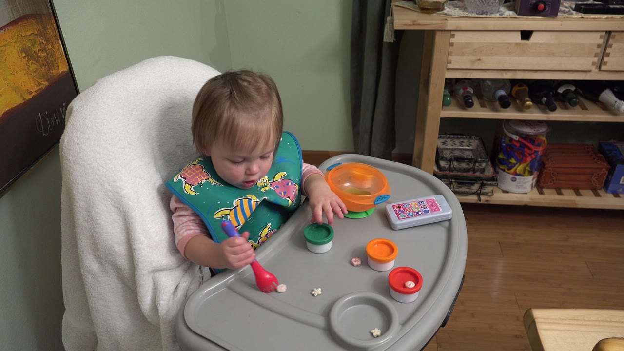 At 0:01, the baby girl remains seated in the high chair in Westminster, Colorado, with one hand near a green-lidded container and the red play tool on the tray.