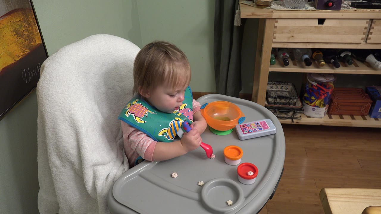 Toddler in a high chair holding a red spoon beside a bowl and small food pieces.