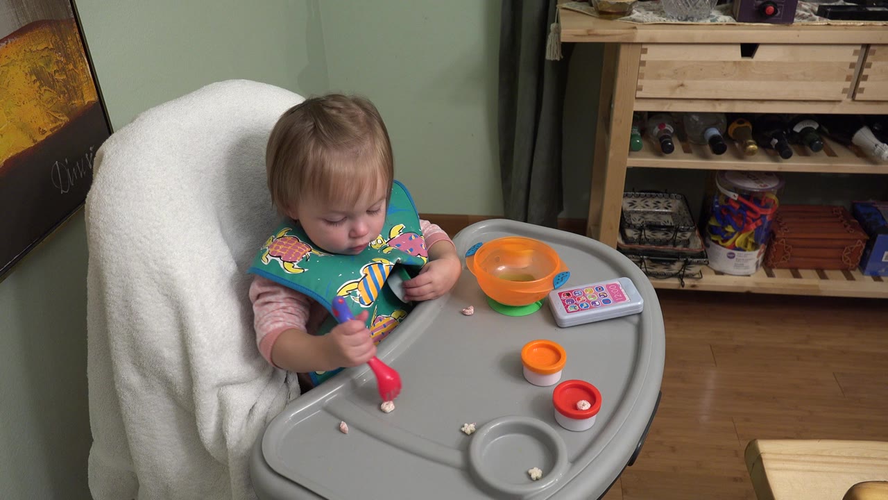 At 0:01 in Westminster, Colorado, the baby keeps their gaze on the tray while the red utensil is held near small food pieces beside orange bowls and a toy phone.