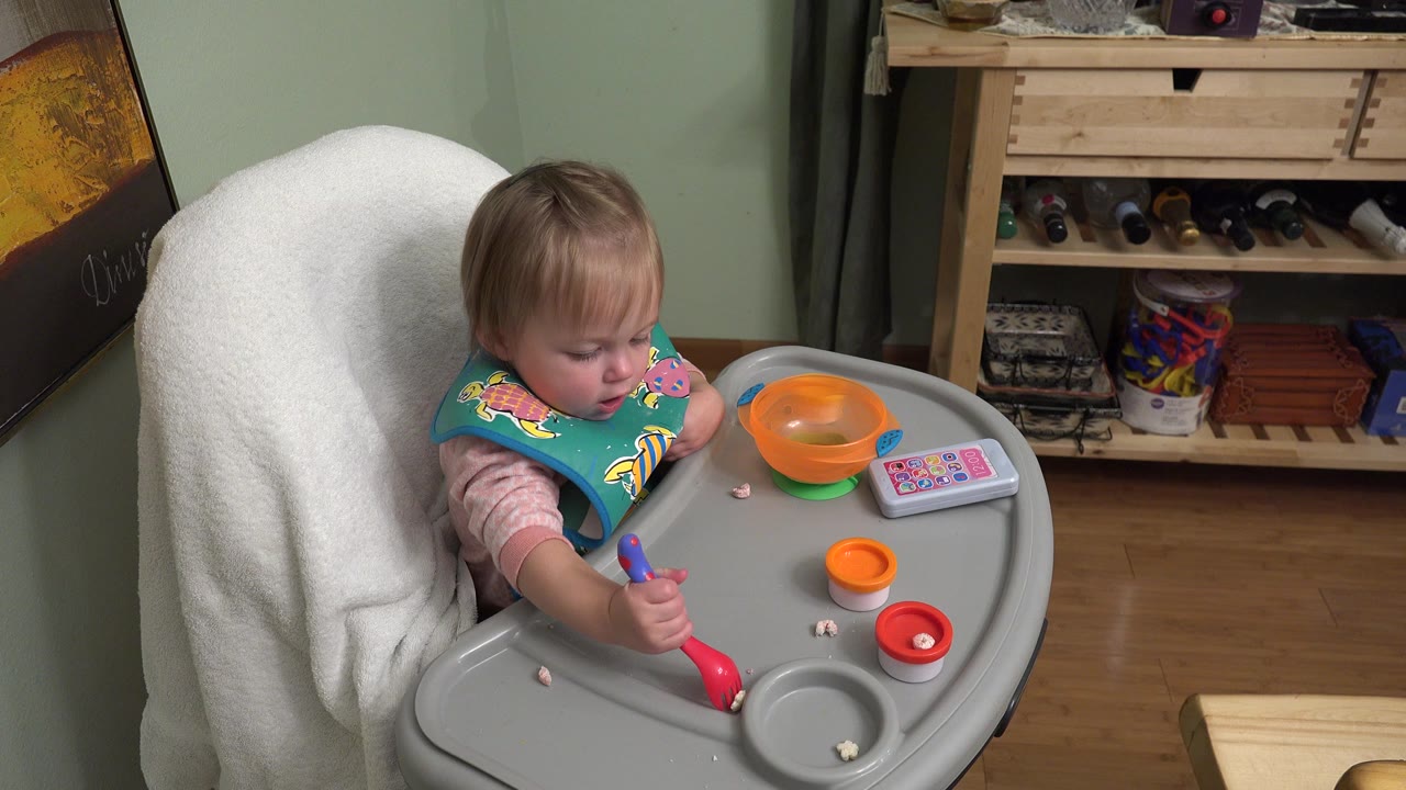 Young child in Westminster, Colorado sits in a high feeding chair with a colorful bib, holding a red and blue spoon near small food pieces on the gray tray.