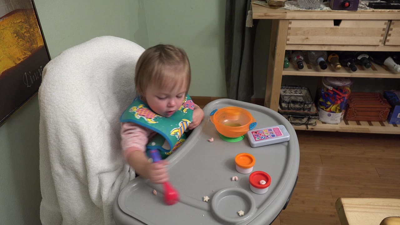 At 0:02, the child in Westminster, Colorado reaches the spoon farther across the gray tray, with the orange bowl and red cup-like toy still on the right side.
