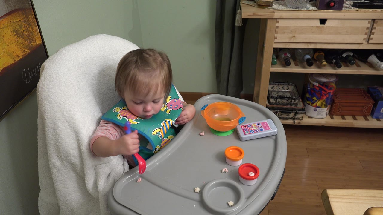The Westminster, Colorado toddler looks down toward small food pieces on the high chair tray while holding the red spoon close to the tray surface.