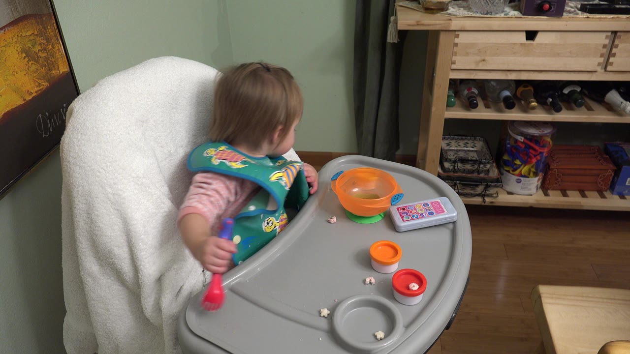 Baby in a highchair holds a colorful utensil beside bowls and crumbs.