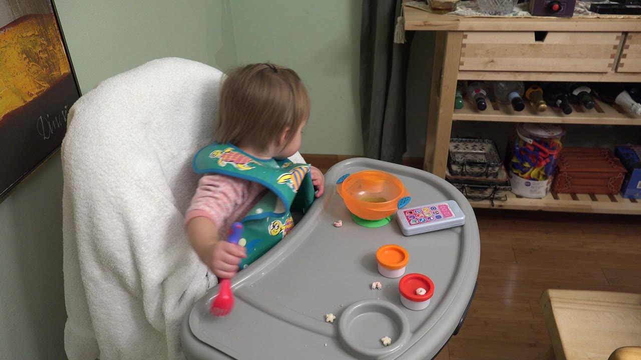 At 0:01 in Westminster, Colorado, the baby remains turned toward the left side of the room while the highchair tray holds an orange bowl, a toy phone, crumbs, and small containers.