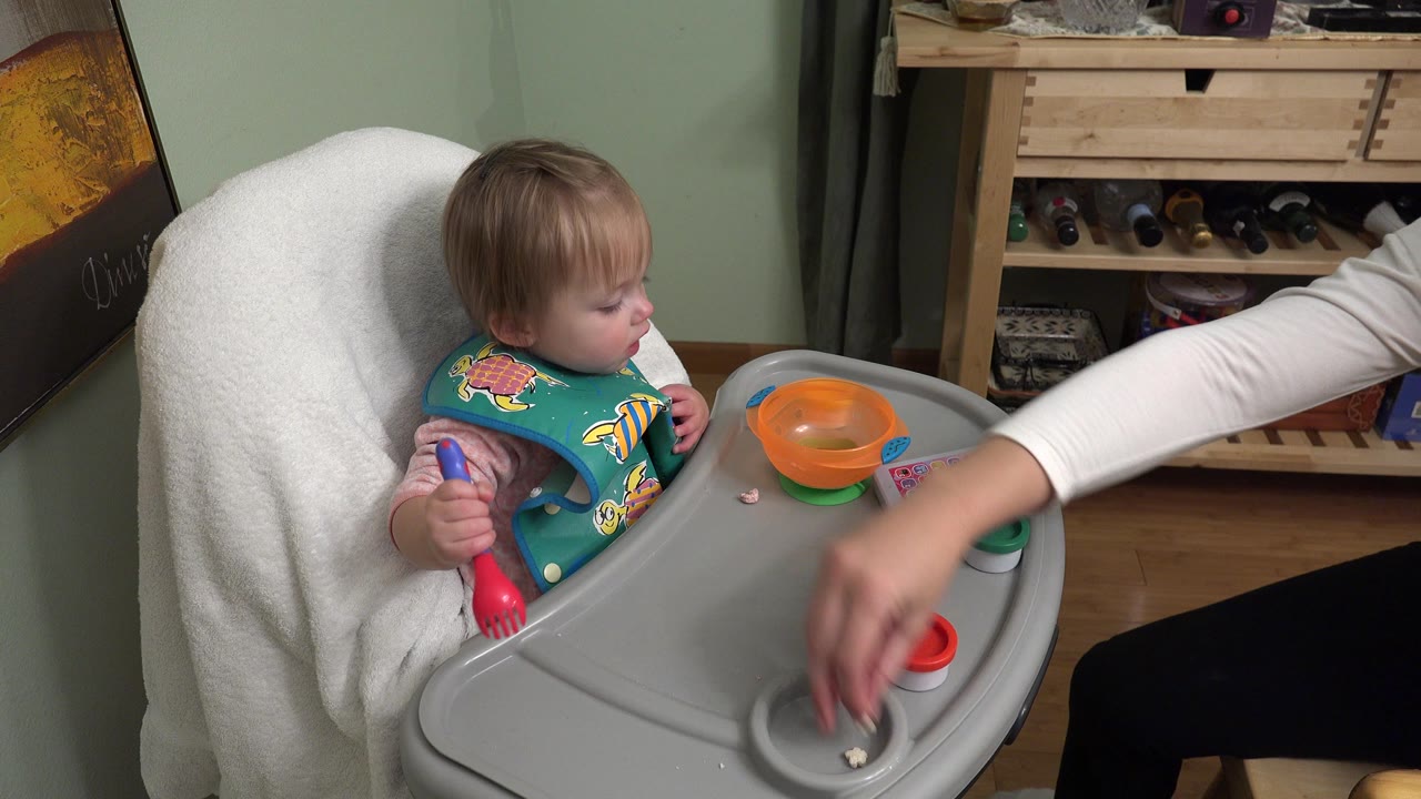 In Westminster, Colorado, a toddler sits in a gray high chair with an orange bowl nearby while an adult hand reaches across the tray toward small snack containers.
