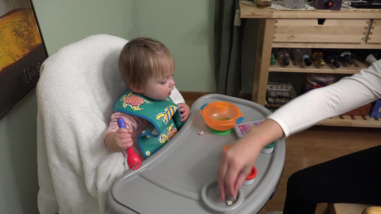 In Westminster, Colorado, the toddler holds a red and blue spoon in the high chair as an adult hand in a white sleeve touches a small container on the tray.