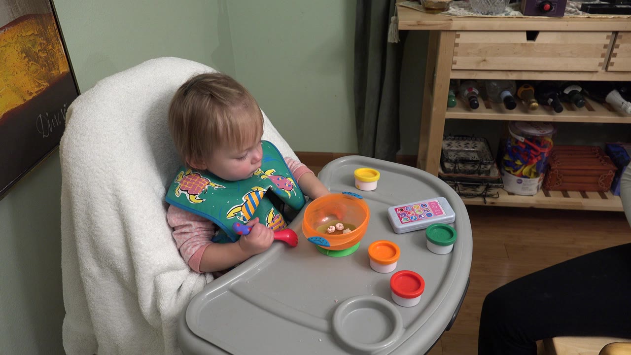 At 0:01 in Westminster, Colorado, the toddler looks toward the orange bowl while holding a spoon, with the cup and toy phone still arranged on the gray tray.