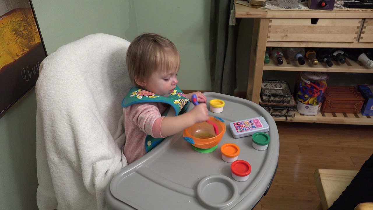 At 0:01, the child leans toward the tray with a utensil in hand, with yellow, green, orange, and red toy containers arranged around the bowl.