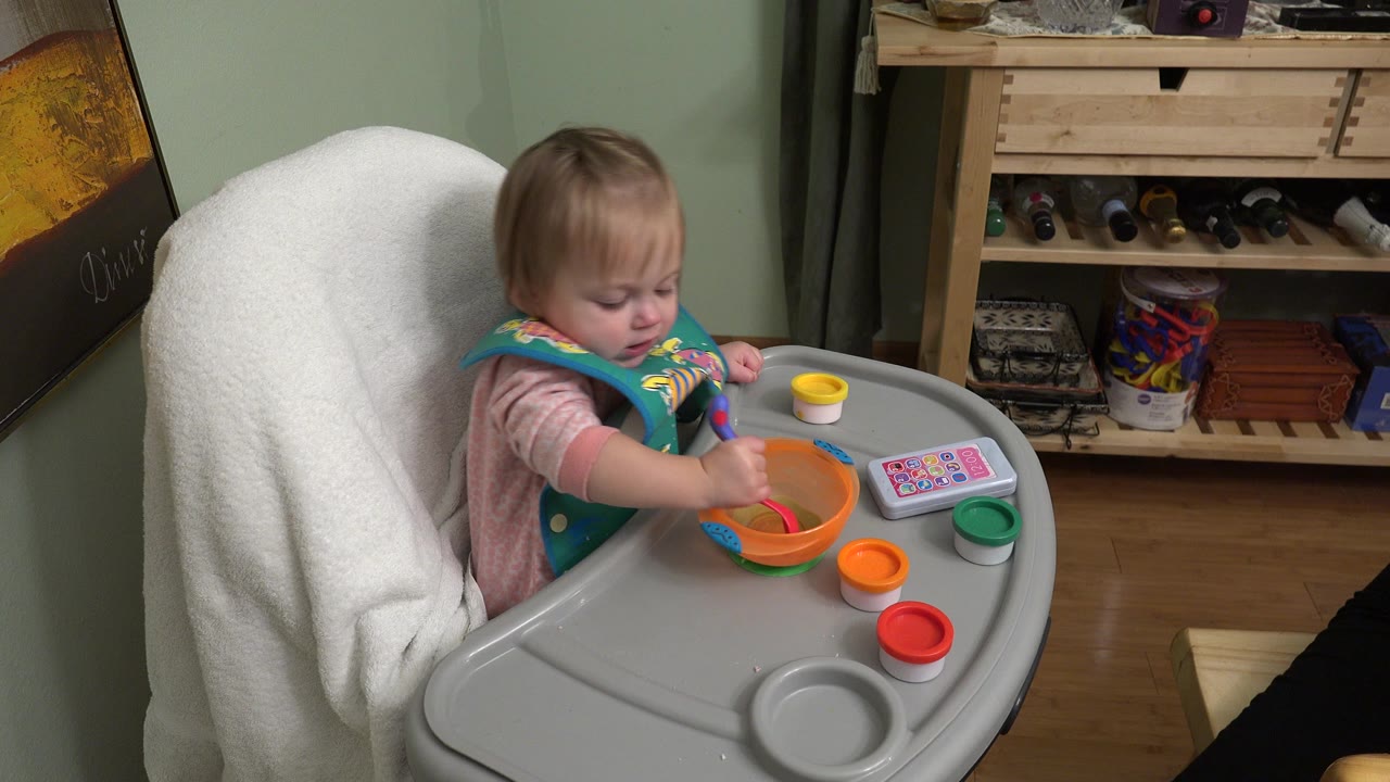 Baby in a high chair reaches a spoon into an orange bowl.