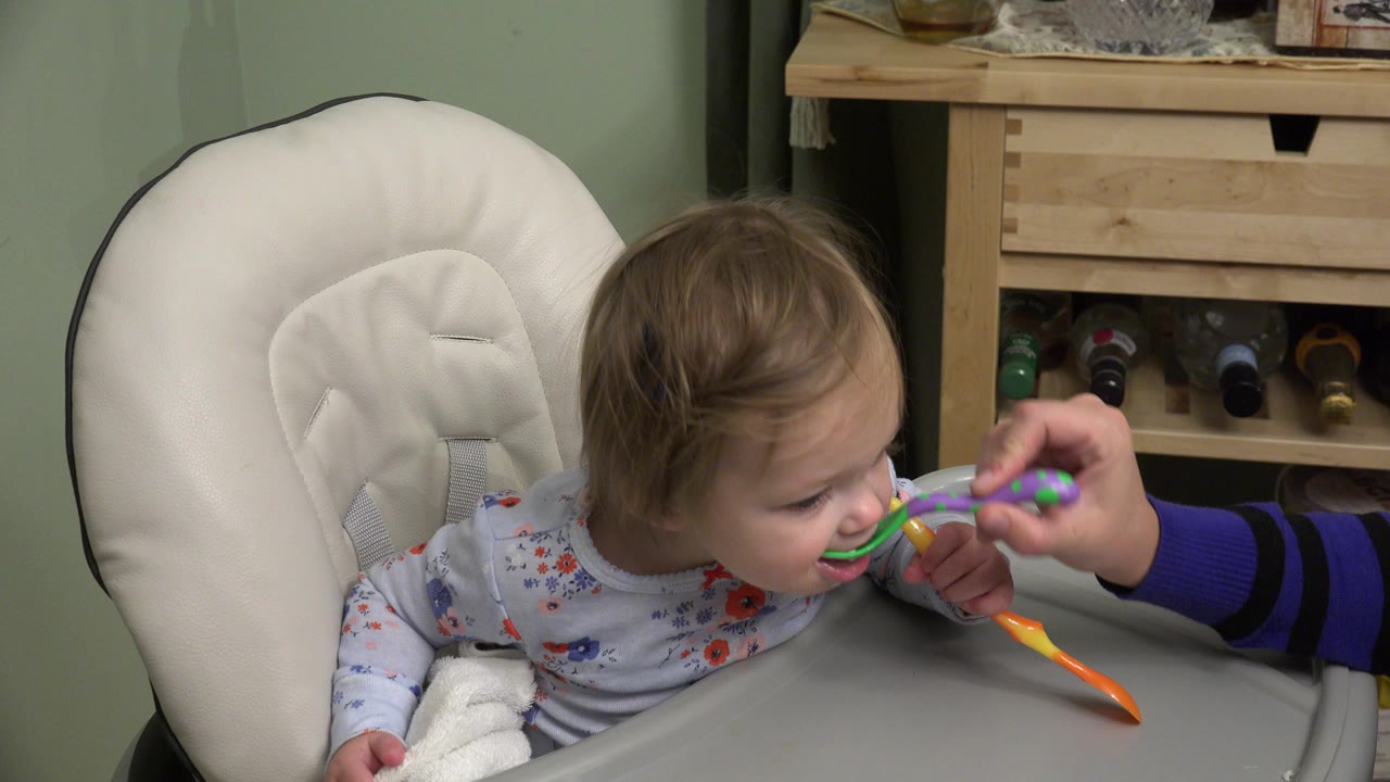 Baby in highchair being fed with a spoon while holding another spoon.