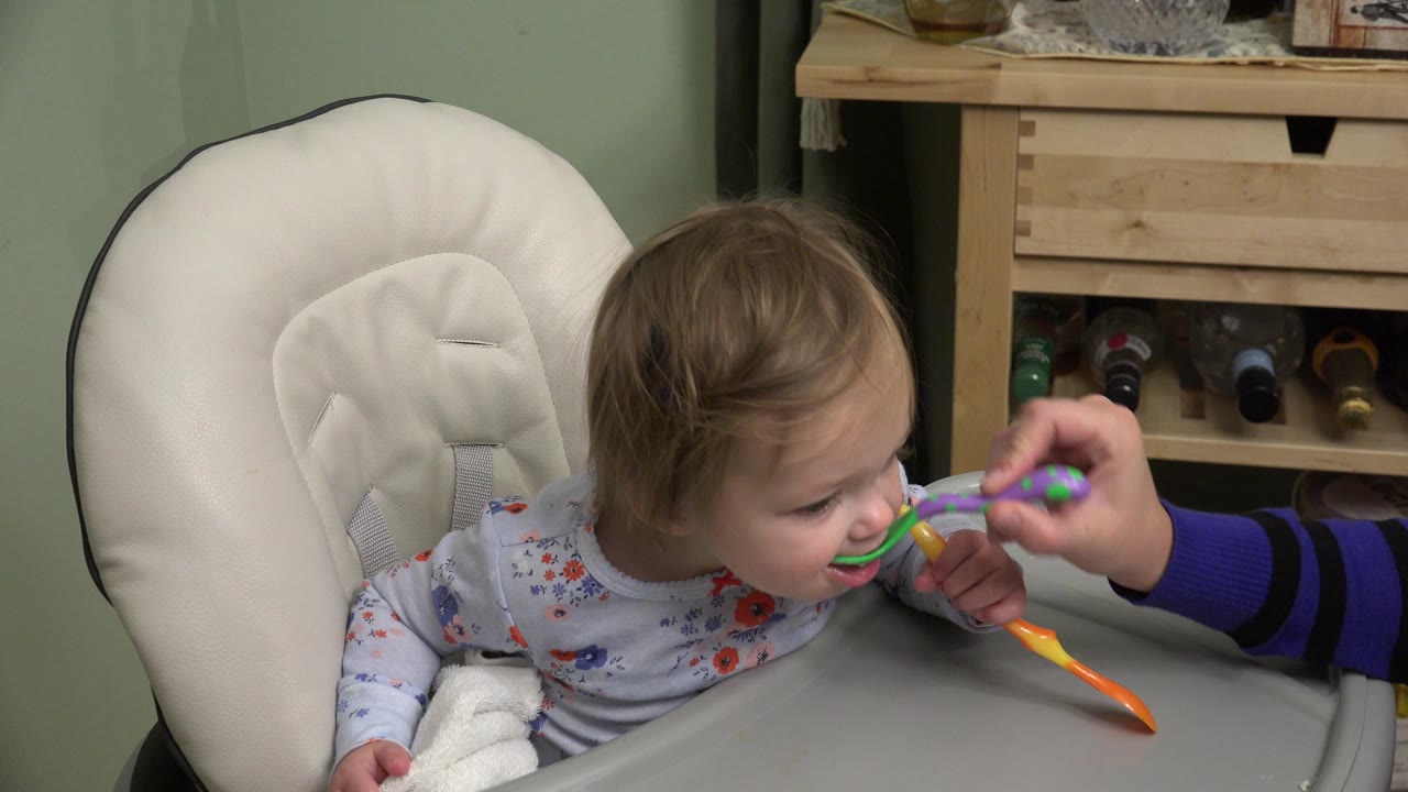 In Westminster, Colorado, the adult hand holds the green spoon at the baby's mouth while the baby leans slightly right and keeps the orange spoon upright in one hand.