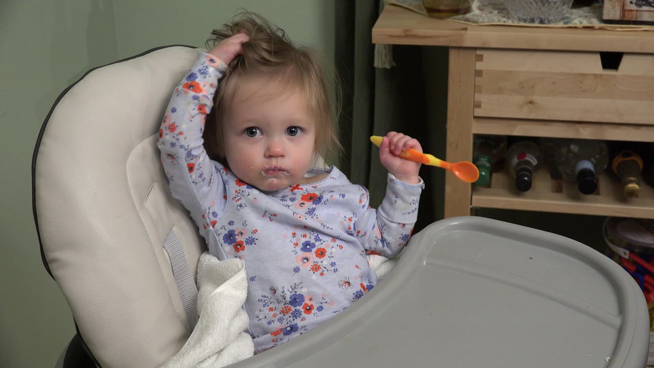 Toddler in a highchair holding an orange spoon and touching her hair.