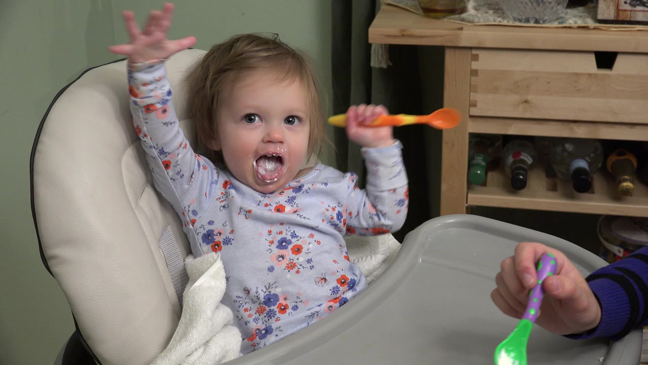 Toddler in a booster seat raises one hand while holding a spoon.