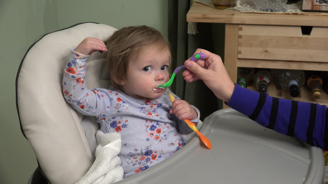 The baby remains seated in the high chair in Westminster, Colorado with food around the mouth as the adult's striped sleeve and green spoon stay near the face.