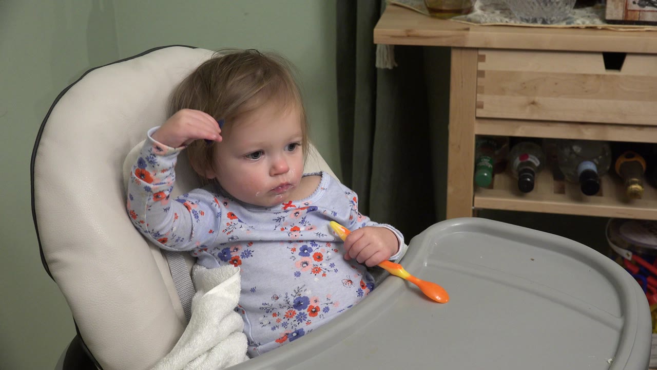 Baby in a high chair touches hair while holding a spoon.