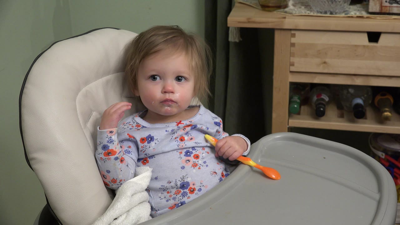 Baby seated in a booster seat holding an orange spoon.